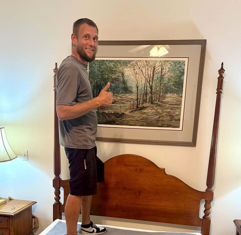 Beach House Moving owner Les hanging a framed landscape above a four-poster bed at a Lily Pads Interiors estate sale in Niceville, FL